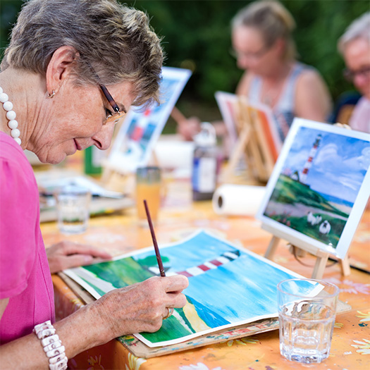 An older woman painting at an outdoor table.